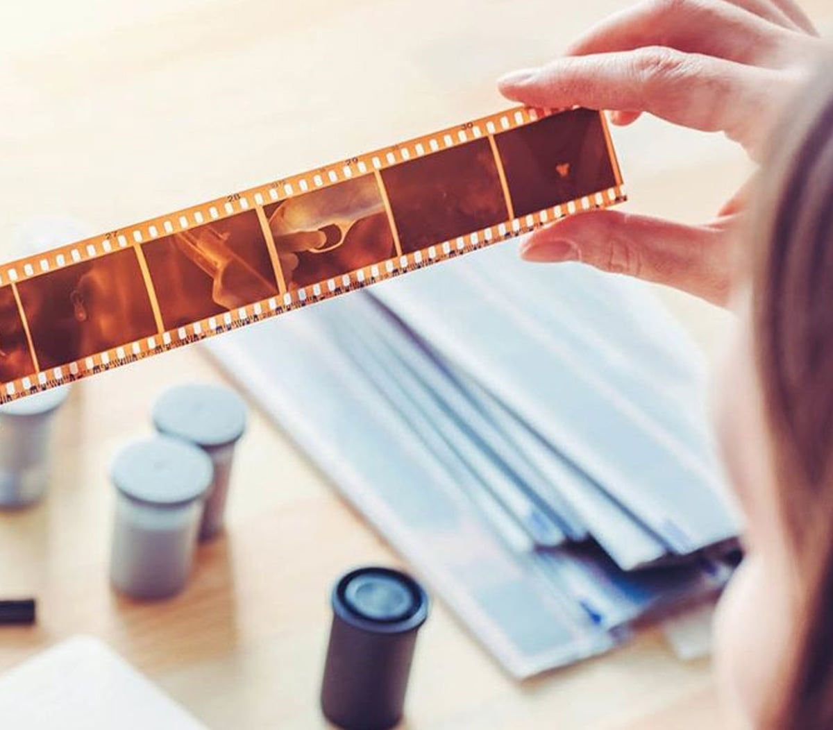 A person holds a strip of photographic film negatives up to the light, with film canisters and photo envelopes on a table in the background.