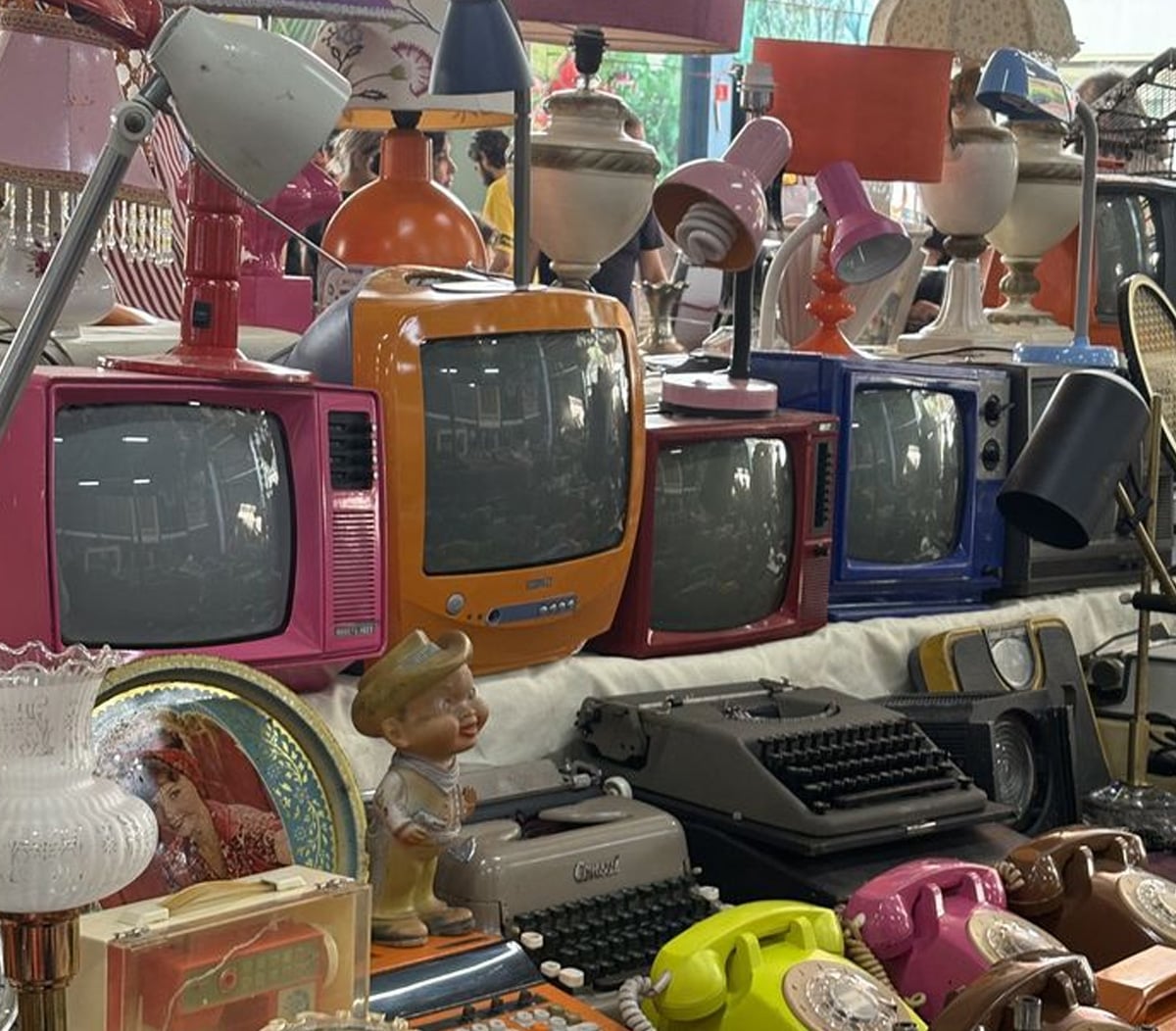 A table at a flea market displays colorful vintage TVs, retro lamps, old telephones, a typewriter, and decorative items, with people browsing in the background.
