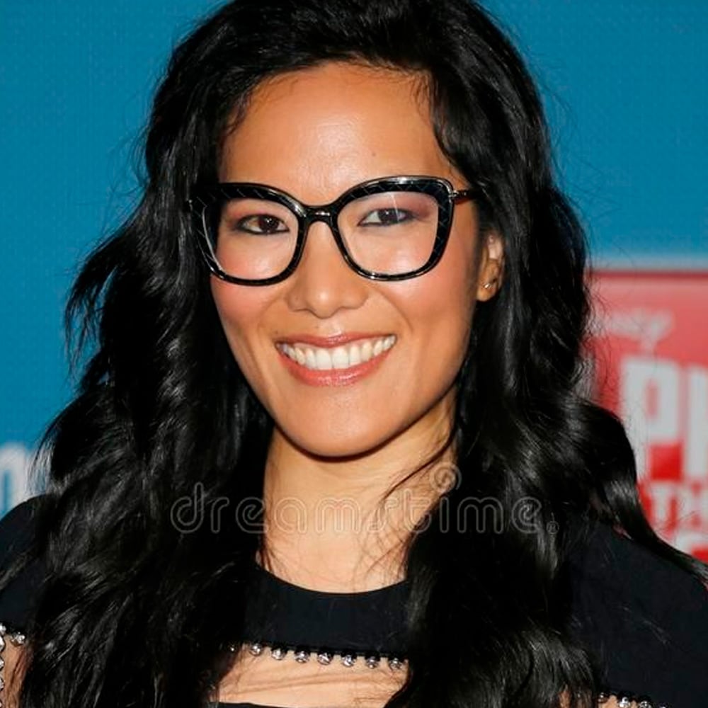 A woman with long, wavy black hair and black glasses smiles at the camera in front of a blue background. She is wearing a black top with sheer details.