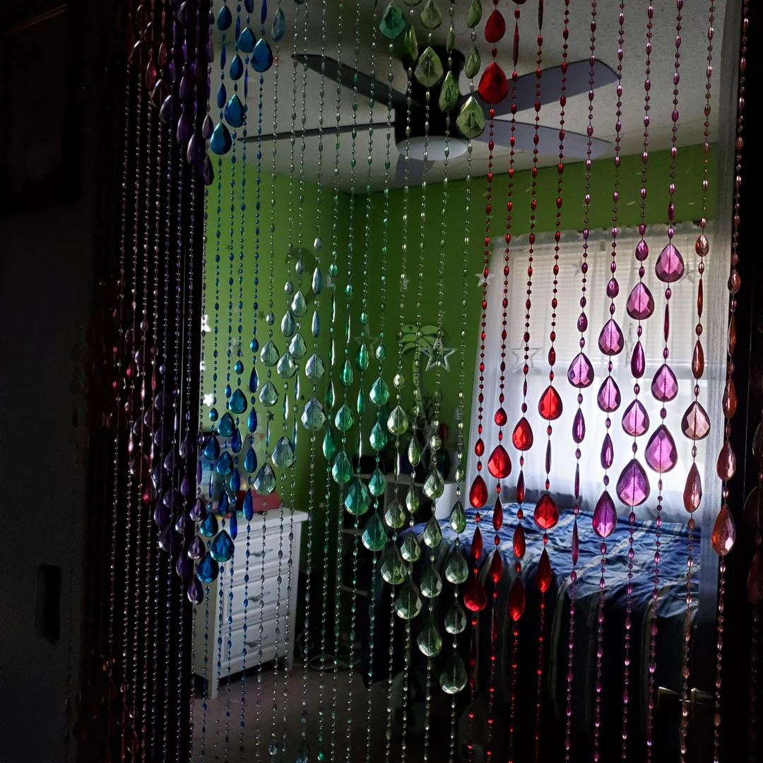 Colorful beaded curtains in purple, blue, green, and red hang in front of a bedroom with green walls, a ceiling fan, a bed, and a small white dresser near a window with sheer curtains.