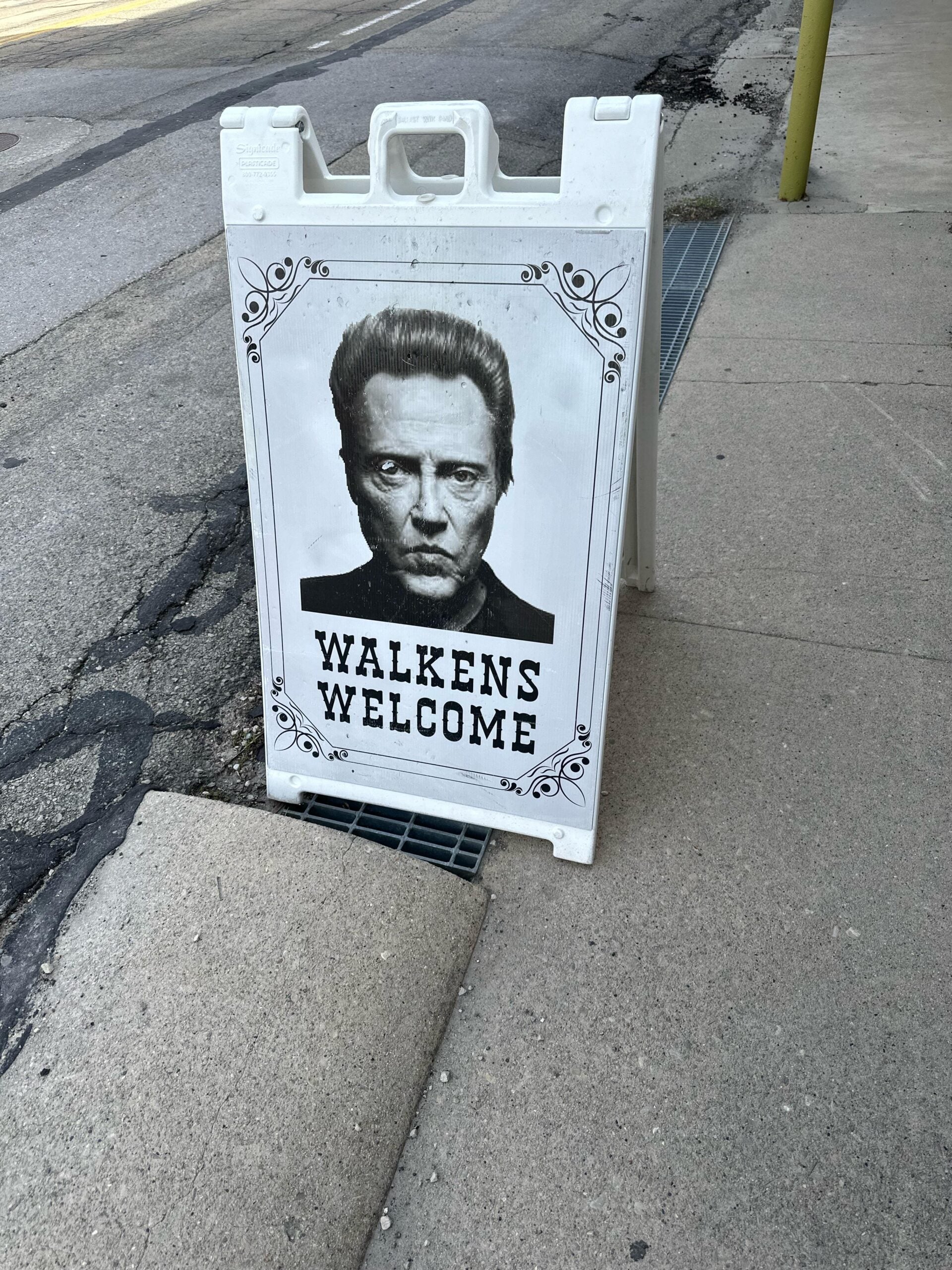 A sidewalk sign with a black-and-white illustration of a man's face and the words "WALKENS WELCOME" underneath, positioned near the edge of a street and sidewalk.