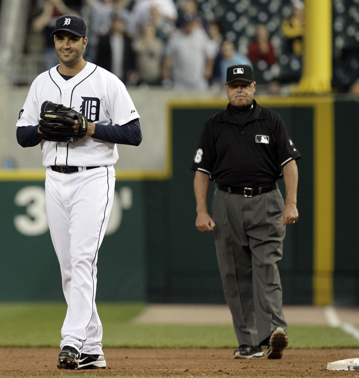 A baseball player in a white Detroit Tigers uniform stands on the field smiling, holding a glove. An umpire in a black uniform stands in the background, and blurred spectators are visible in the stands.