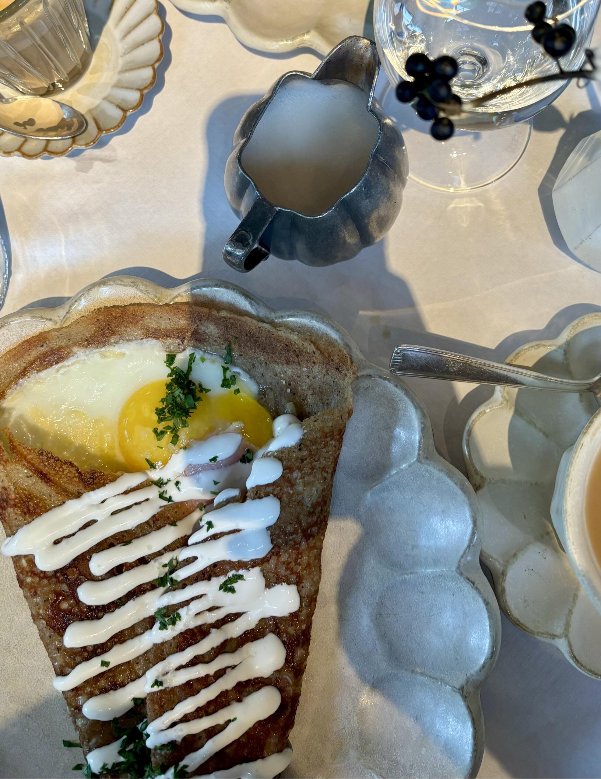 A savory crepe topped with a sunny-side-up egg, chopped herbs, and drizzled white sauce sits on a scalloped plate. Nearby are a cup of tea, a small metal pitcher, glassware, and candles on a white tablecloth.