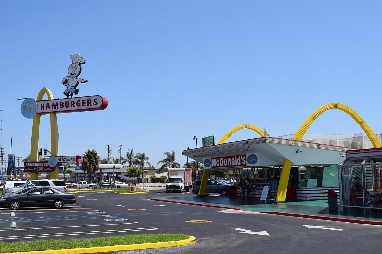 A vintage McDonald’s restaurant with golden arches, retro signage, and a large parking lot under a clear blue sky. A tall sign features a cartoon chef atop the McDonald’s name and the classic “hamburgers” text.