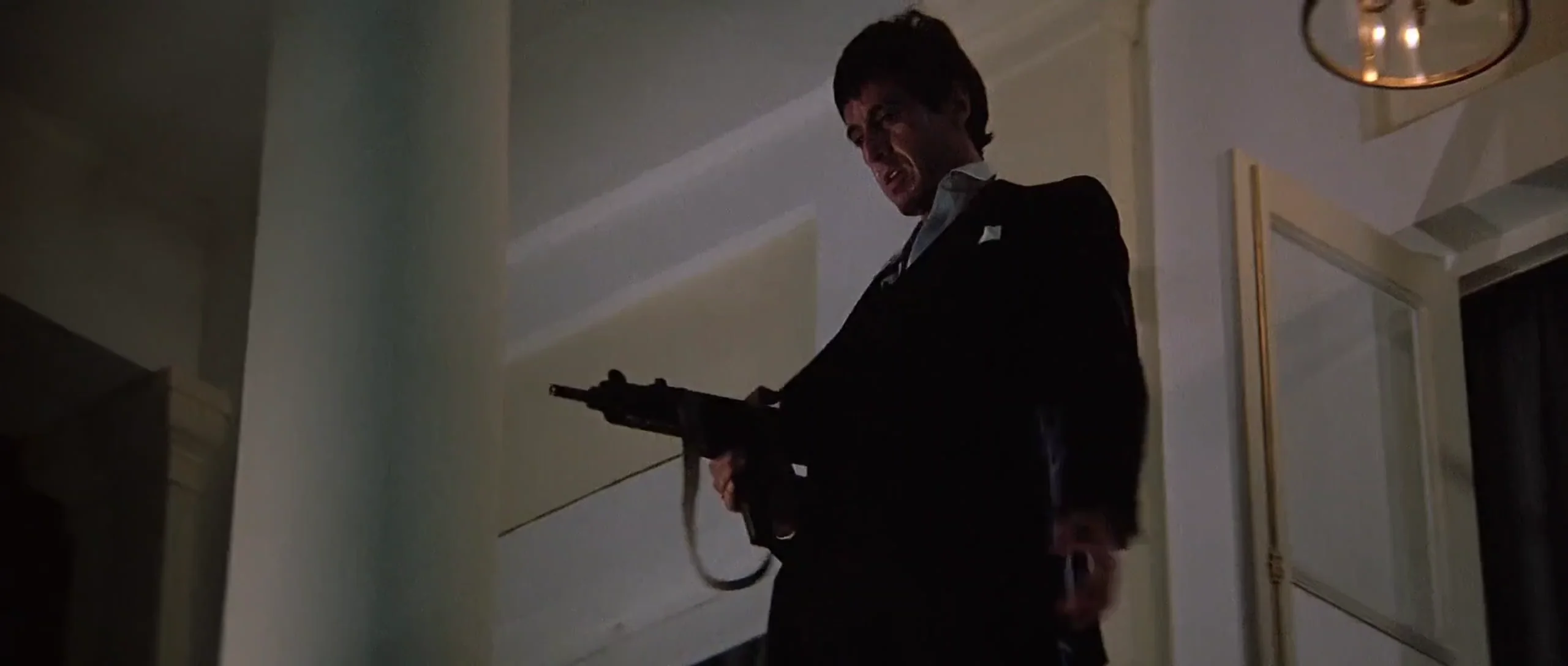 A man in a dark suit stands indoors, holding a gun and looking down with an intense expression. The scene is dimly lit, with light coming from above, creating dramatic shadows on the walls.