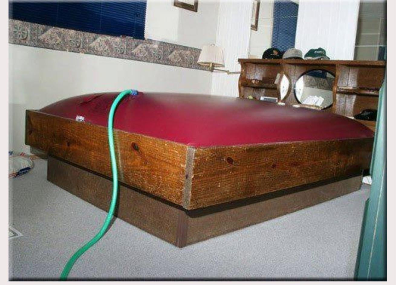 A wooden waterbed frame filled to overcapacity, with the red mattress bulging upward and a green hose attached, causing the bed to appear swollen and stretched.
