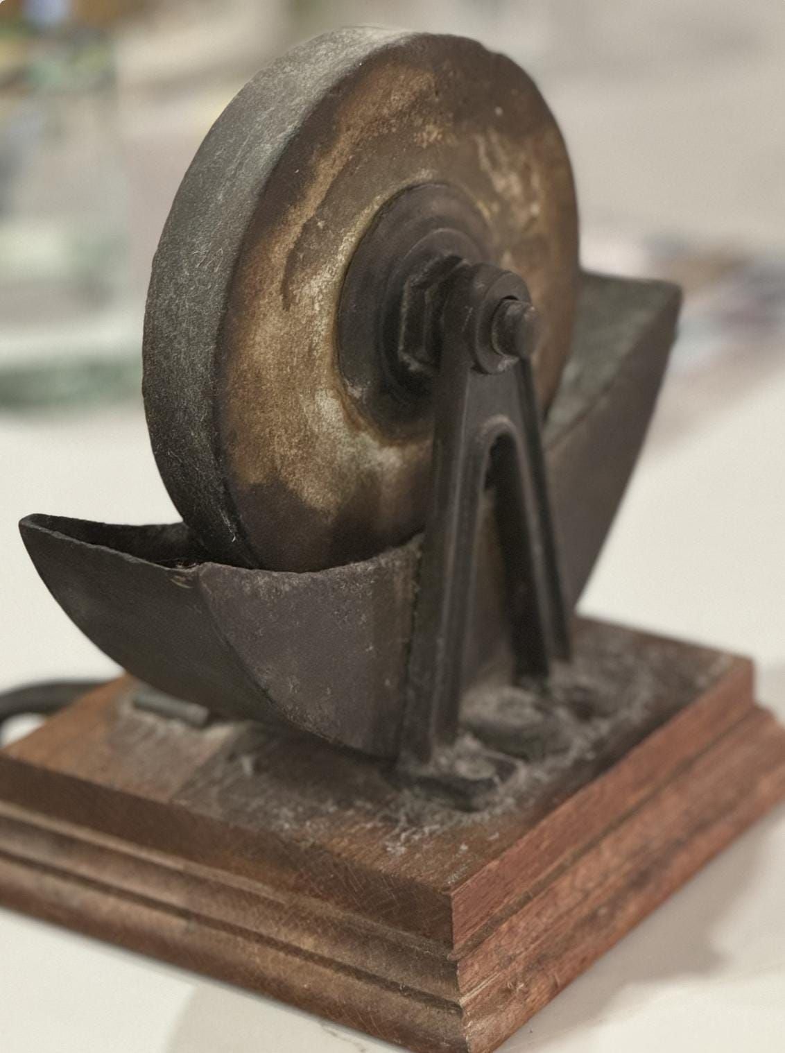 A close-up of a metal sculpture resembling a grinding wheel mounted on a wooden base, with geometric metal supports and a weathered, rustic appearance.