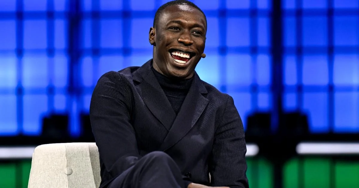 A man in a dark suit sits on a light chair, smiling widely and looking to the side. The background features a blue, grid-like pattern with bright lighting.