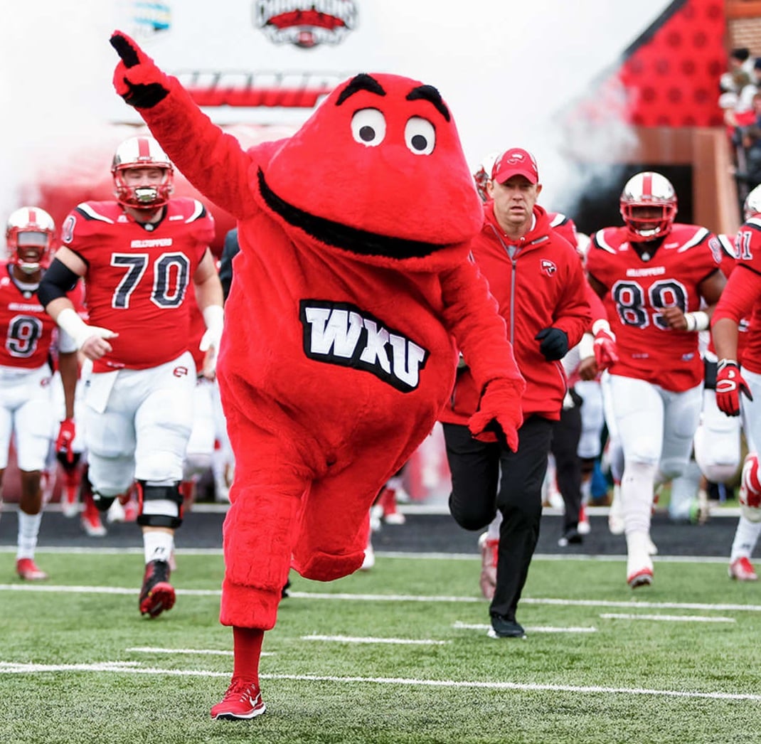 The WKU Big Red mascot leads the Western Kentucky University football team onto the field, with players and a coach running behind, in a stadium setting filled with excitement.