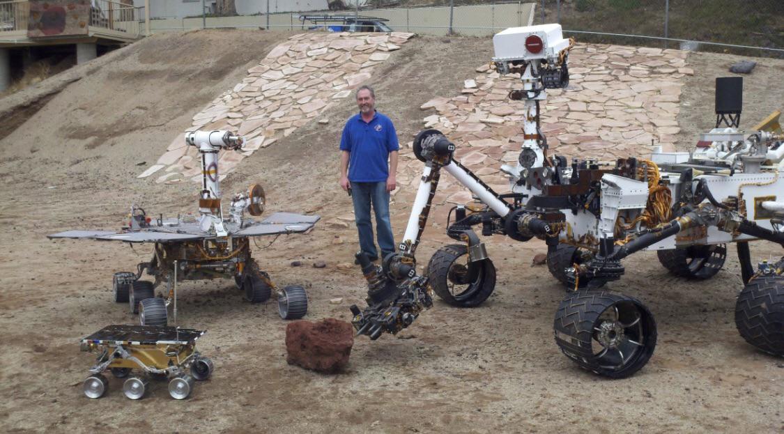 A man stands outdoors between three Mars rover models of different sizes, including Sojourner, Spirit/Opportunity, and Curiosity, displayed on rocky ground with a stone-covered slope in the background.
