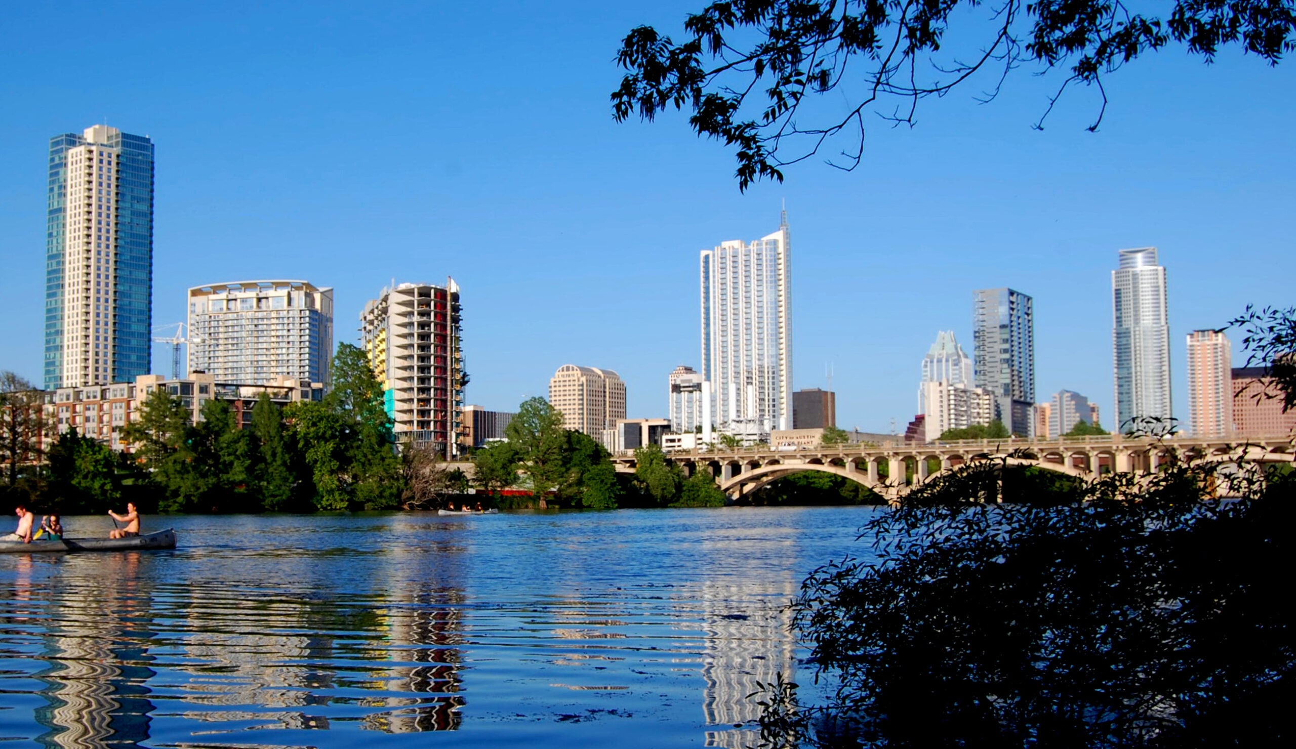 A city skyline with tall buildings and a bridge reflected in a calm river, with trees in the foreground and people paddling a canoe on the water under a clear blue sky.