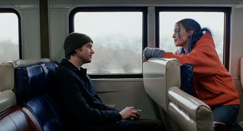 A man in a dark jacket and beanie sits on a train seat, looking at a woman with blue hair and an orange hoodie who is leaning over the back of the seat in front of him, talking animatedly.