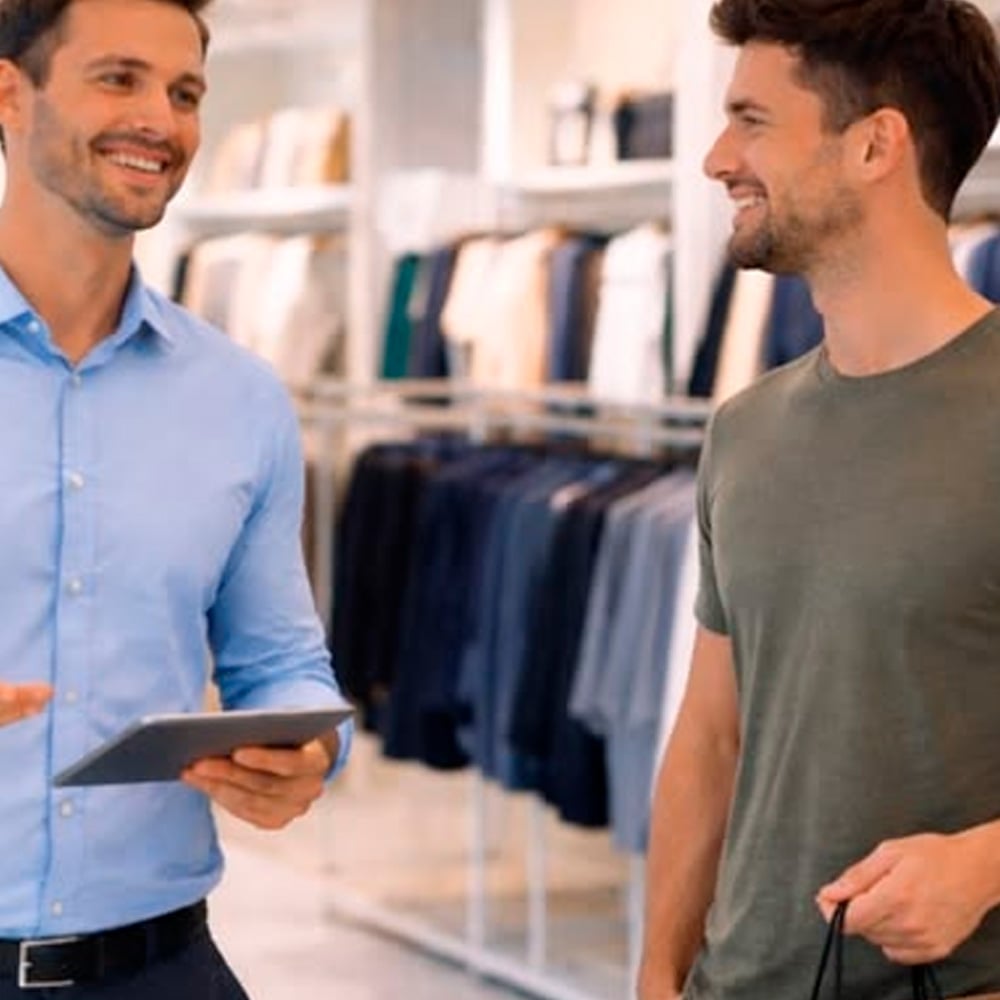 Two men stand in a clothing store, smiling and talking. One holds a tablet and wears a blue shirt, while the other wears a green t-shirt and holds a shopping bag. Racks of clothes are visible in the background.