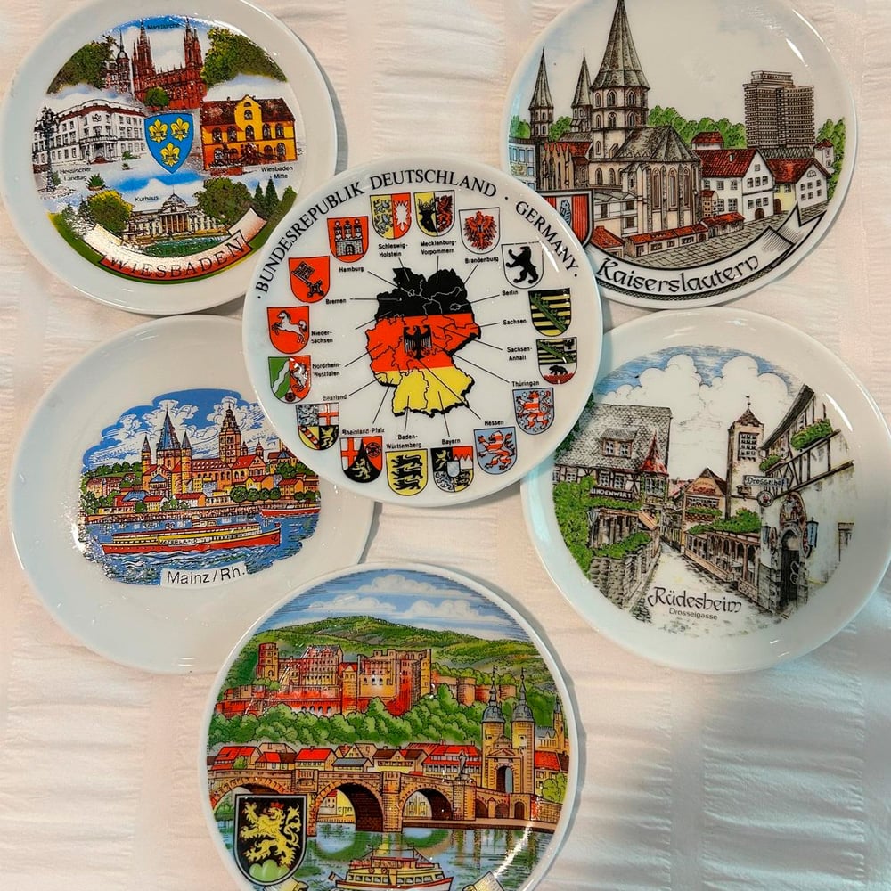 Six decorative plates with colorful illustrations of German landmarks and cityscapes, including Kaiserslautern, Mainz, and Rüdesheim. The center plate features a map of Germany with regional crests and the national flag.