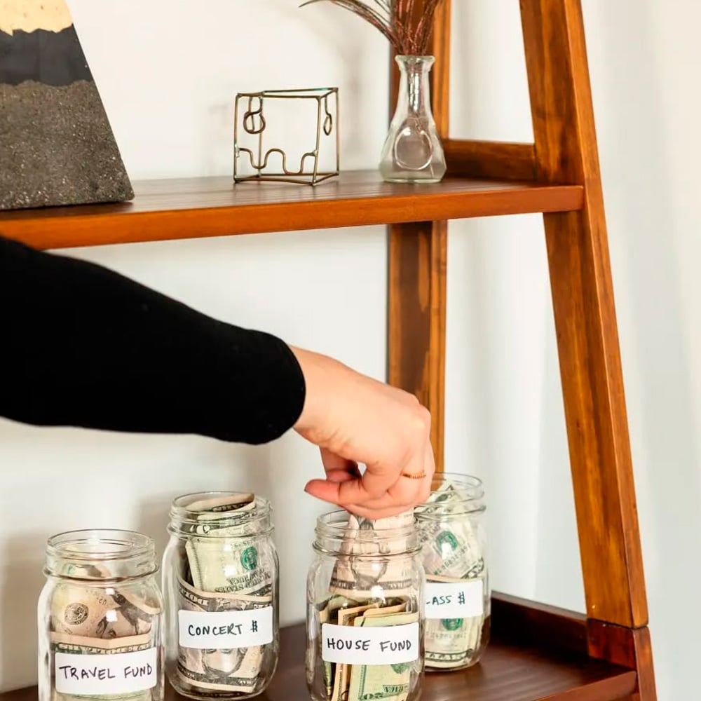 A person places money into a glass jar labeled "House Fund," alongside other jars labeled "Travel Fund" and "Concert," all partly filled with cash, on a wooden shelf.