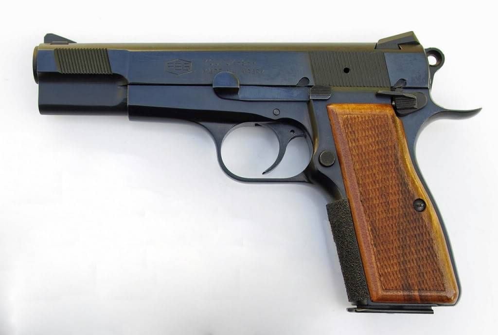 A black semi-automatic handgun with a brown wooden grip is shown on a white background. The pistol's slide and barrel are visible, and the hammer is in the cocked position.