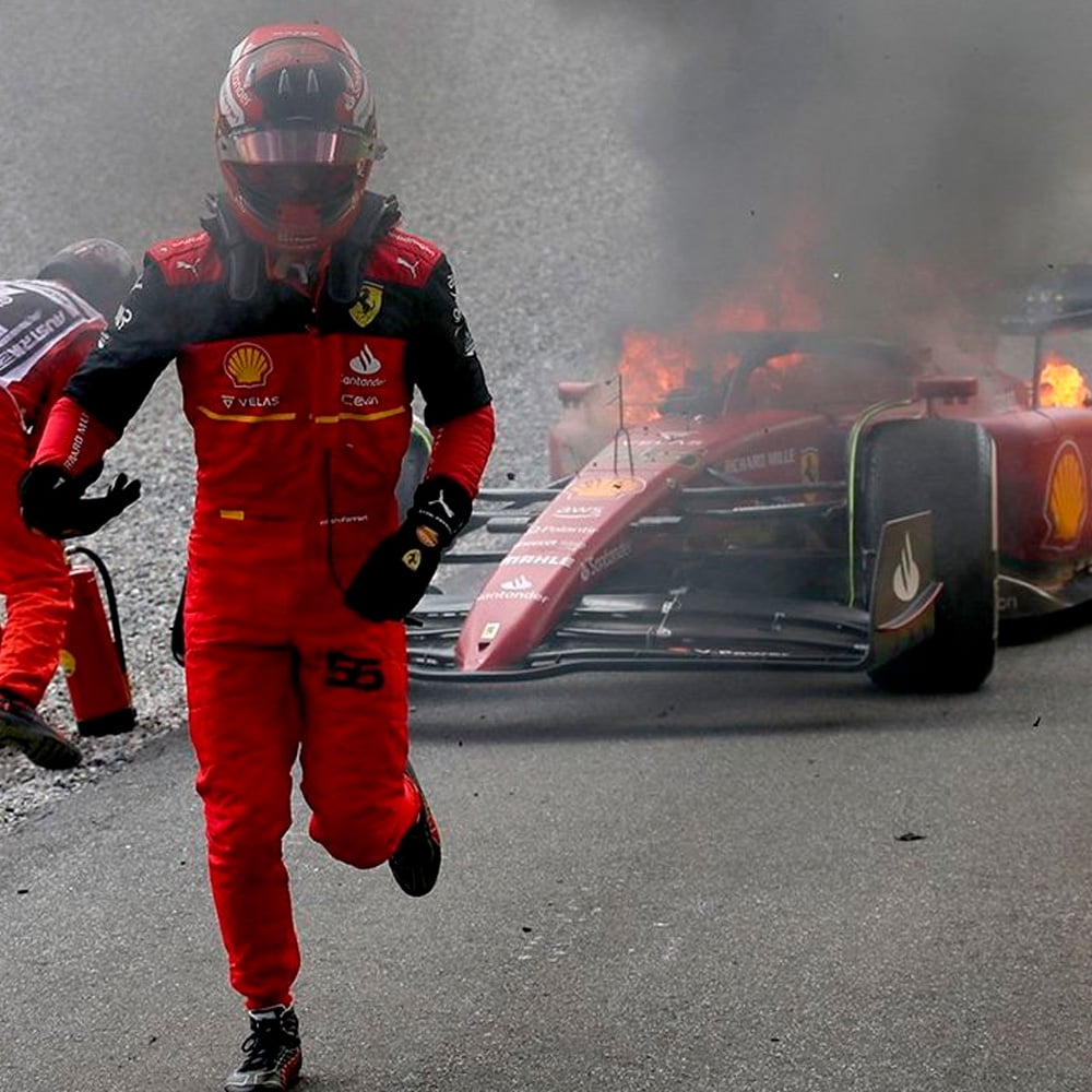 A Formula 1 driver in red Ferrari gear runs away from his race car, which is on fire and producing thick smoke at the back of the vehicle. Another person is visible near the flames in the background.