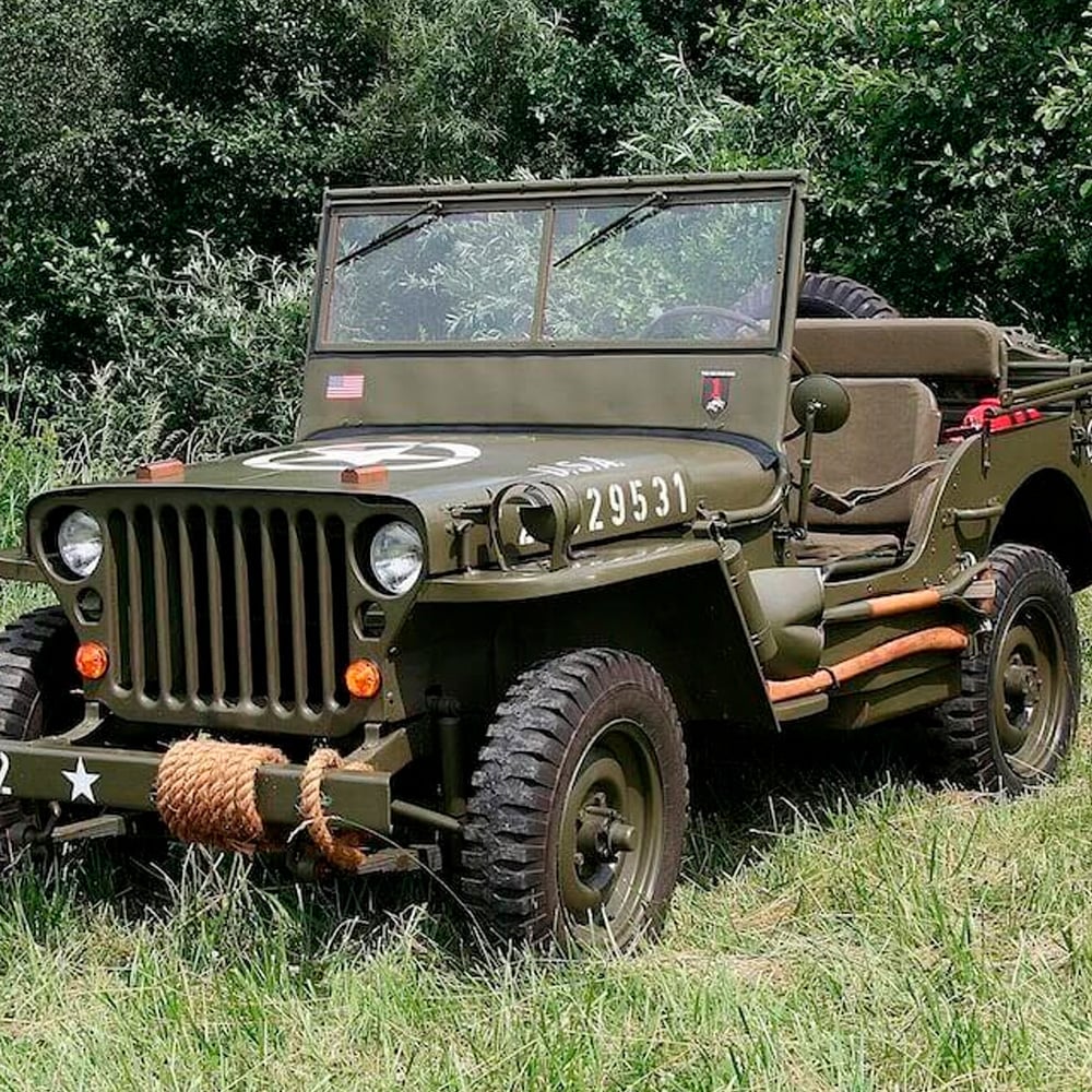 A vintage olive green military jeep with a rope tied to the front bumper is parked on grass, surrounded by trees and bushes. The vehicle has markings, an open top, and minimal seats.