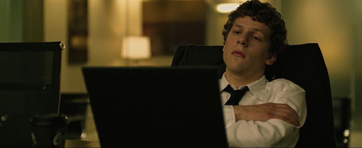 A young man with curly hair, wearing a white shirt and black tie, sits back in an office chair with his arms crossed, looking intently at a laptop screen in a dimly lit modern office.