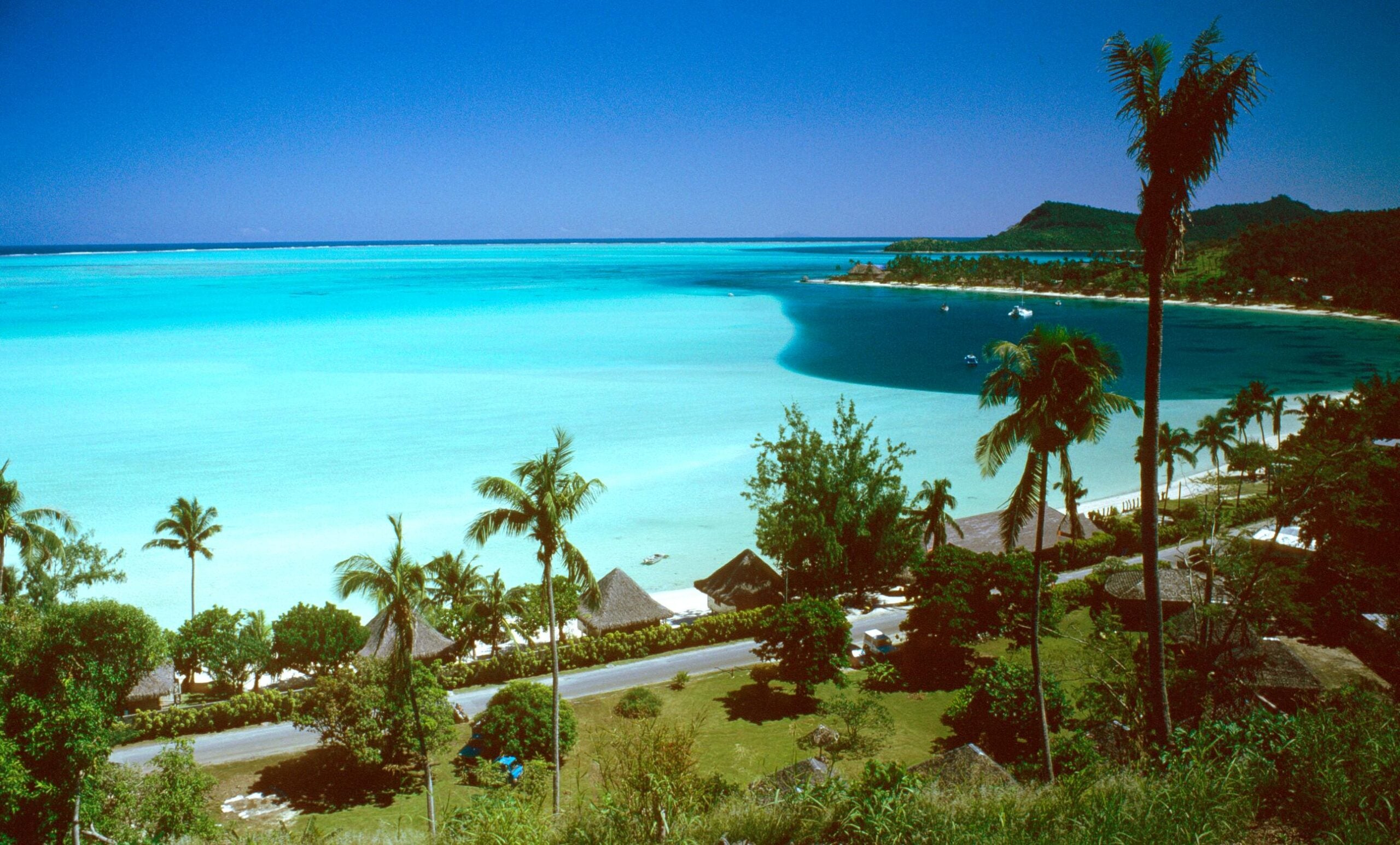 A tropical coastline with turquoise water, lush palm trees, green vegetation, a road, and thatched-roof huts under a clear blue sky.