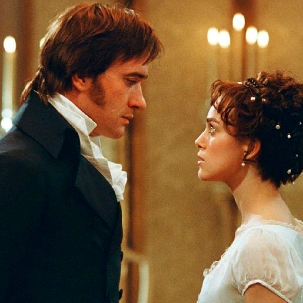 A man and woman in Regency-era clothing stand face to face, gazing at each other intensely, with soft candlelight in the background.