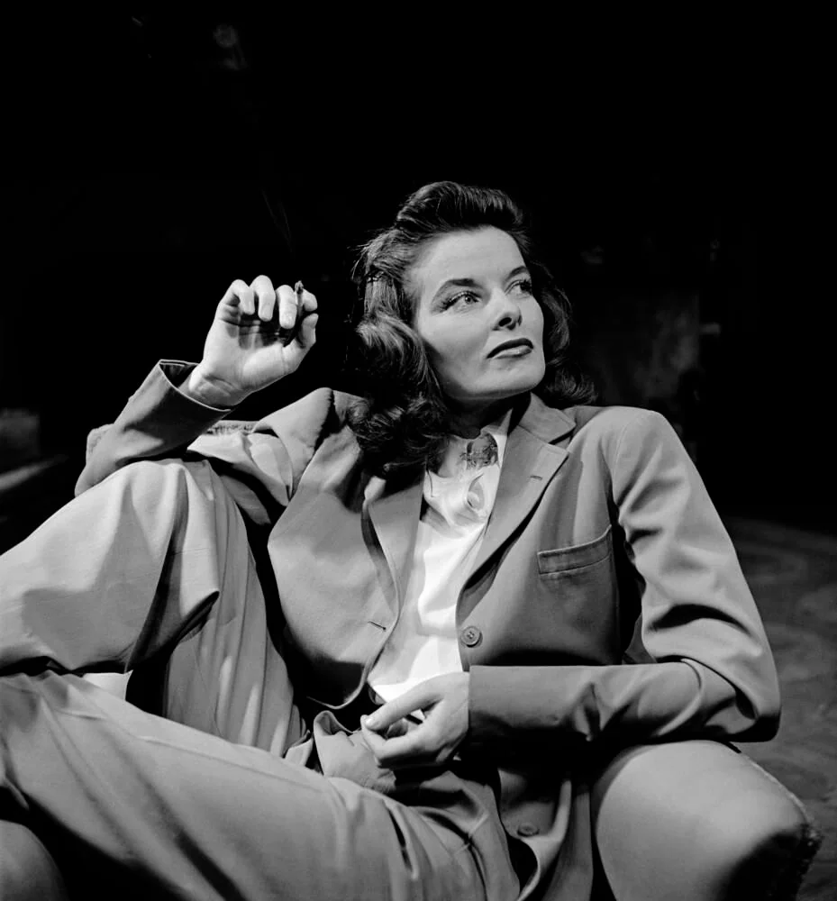 A woman in a suit sits comfortably in a chair, legs crossed, holding a cigarette with a confident expression. The photo is black and white, and the background is dark, drawing focus to her poised demeanor.