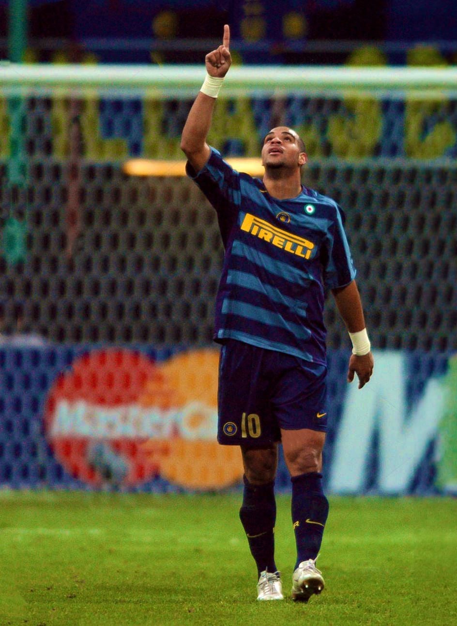 A soccer player in a blue and black striped Inter Milan jersey, number 10, raises his right arm and points upward while standing on the field during a match. The goal net and advertising boards are visible in the background.
