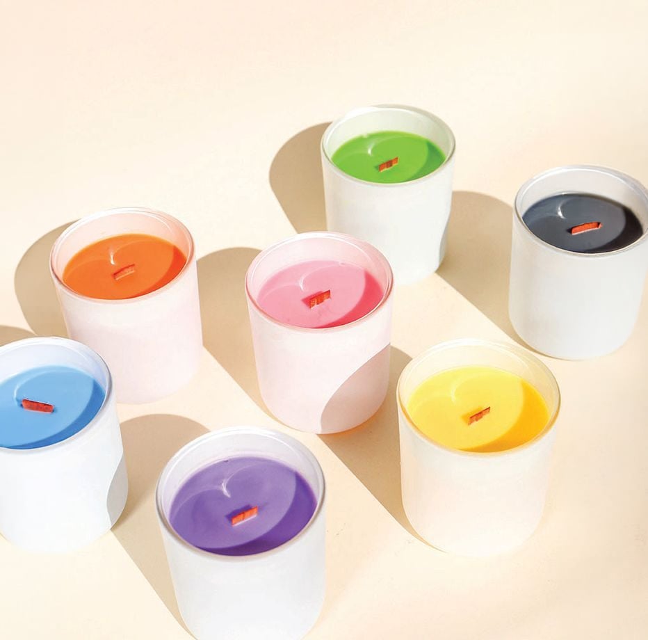 Seven white candle jars with colorful wax—orange, pink, green, blue, yellow, purple, and black—are arranged on a pale surface, each with a wooden wick and casting soft shadows.