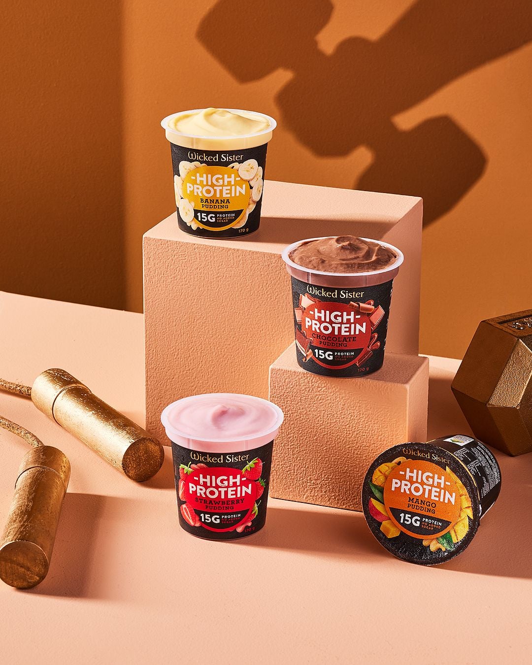 Four Wicked Sister High Protein yogurt cups in mango, chocolate, strawberry, and vanilla flavors are displayed on peach-colored blocks with golden dumbbells in the background against an orange backdrop.