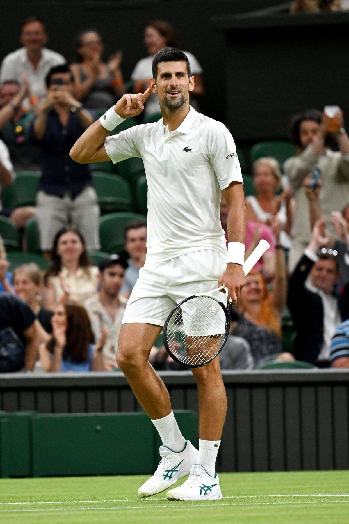 A male tennis player in a white outfit stands on a grass court, holding a racket, and gestures to his ear while smiling. Spectators in the background applaud and cheer.