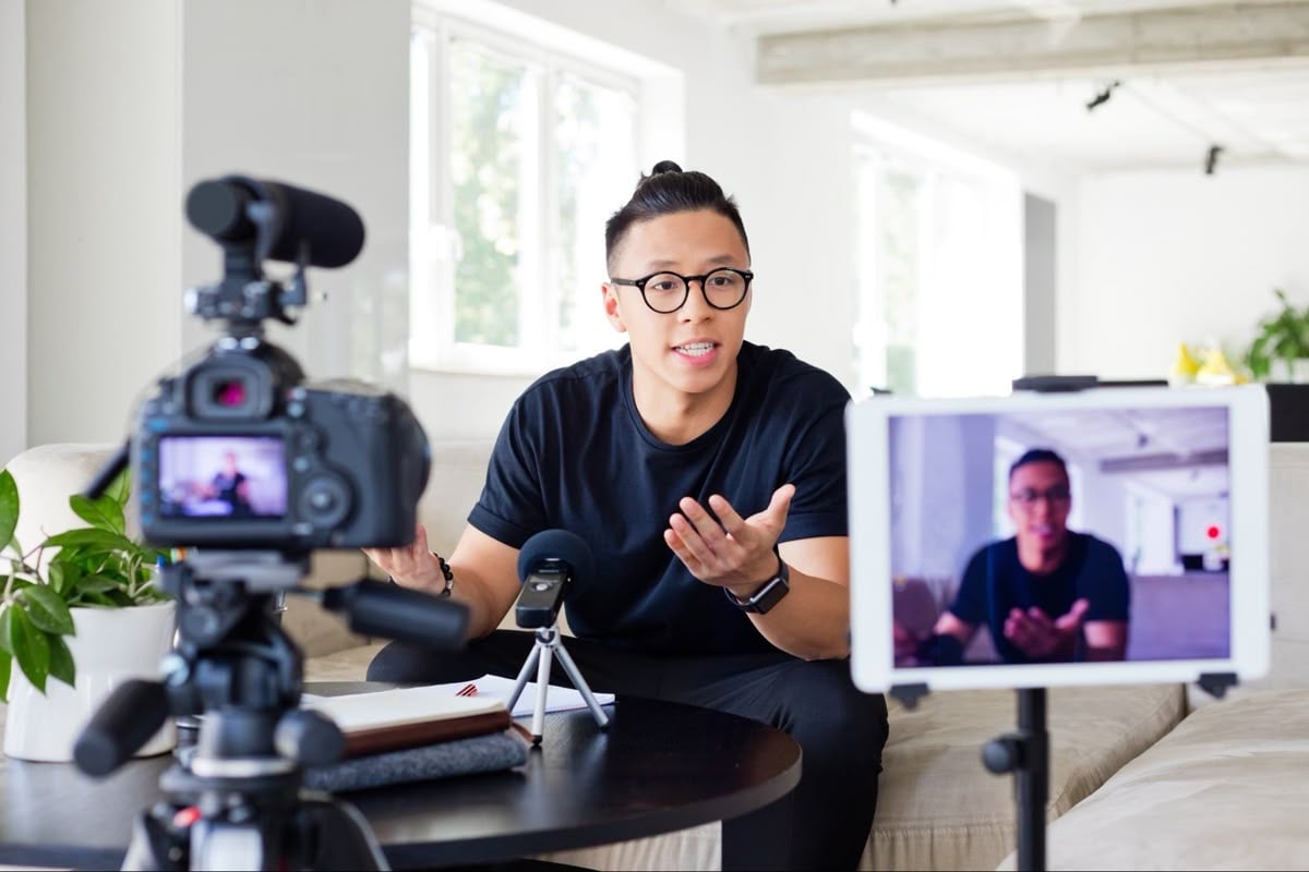 A person wearing glasses sits on a couch, speaking animatedly in front of a camera and microphone, while being recorded. A tablet nearby displays their live image. Bright, modern room in the background.