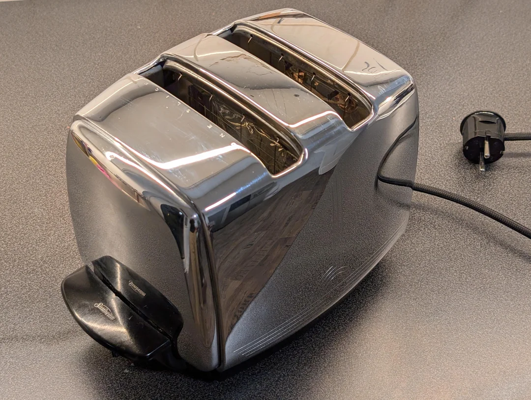 A shiny stainless steel two-slot toaster with a black lever and a power cord on a dark countertop. The toaster's surface reflects part of the surrounding environment.