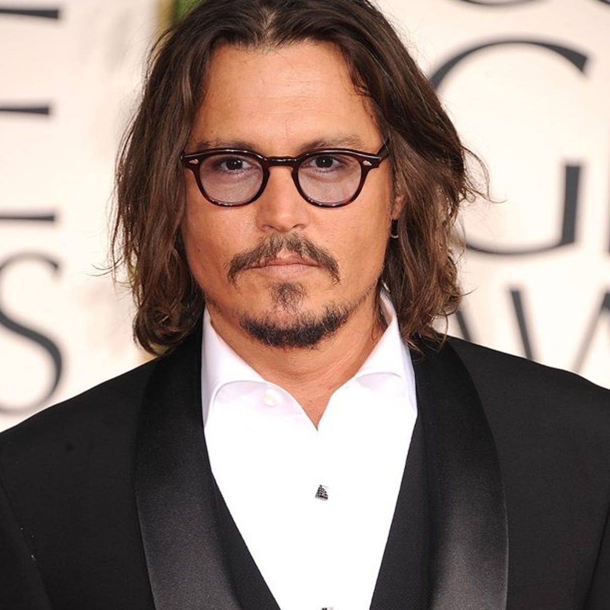 A man with shoulder-length brown hair, a mustache, goatee, and round glasses is wearing a white shirt and black tuxedo jacket, standing in front of a blurred background with large letters.