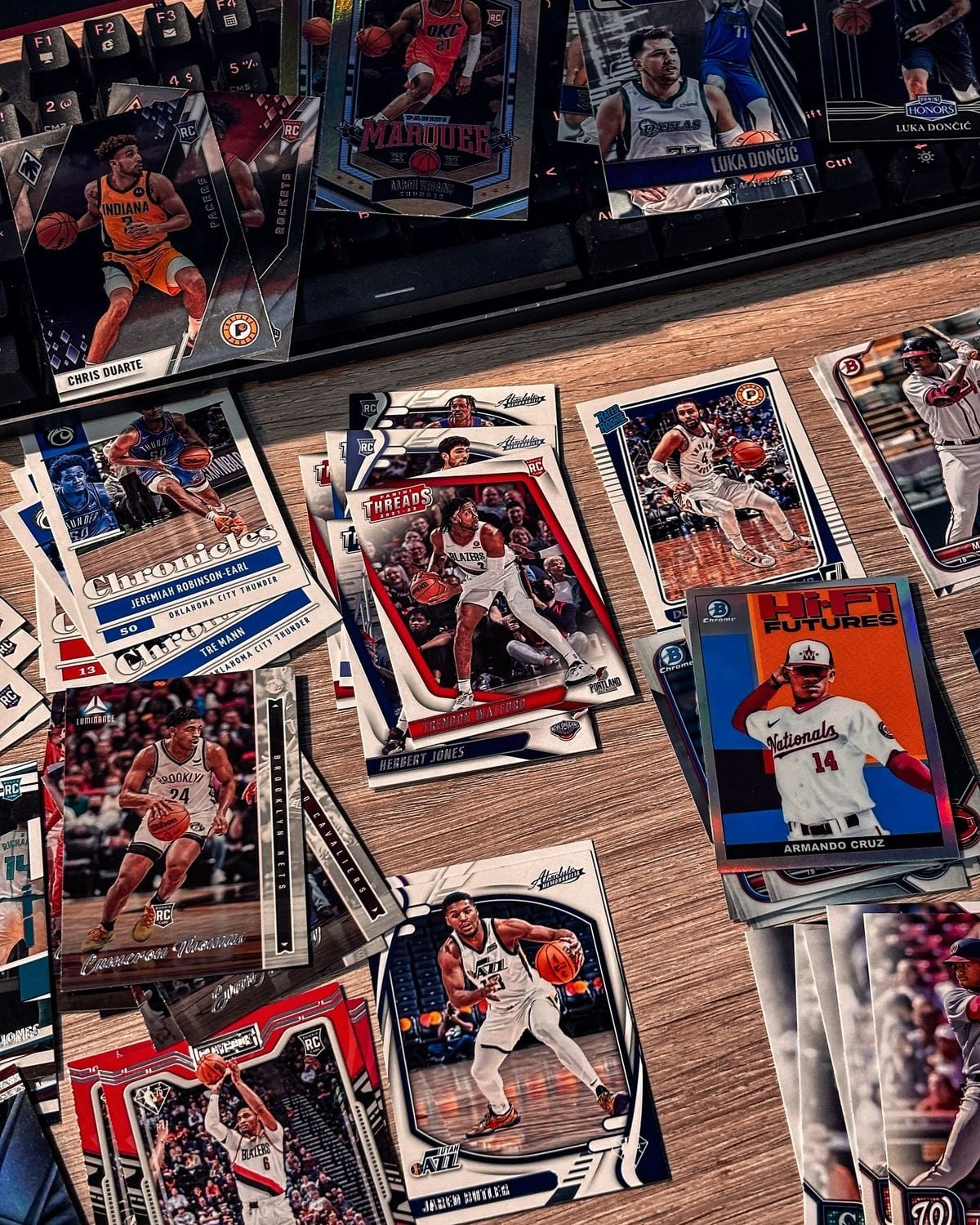 A collection of assorted basketball trading cards is spread out on a wooden desk near a computer keyboard, featuring various players in action poses and different card designs.