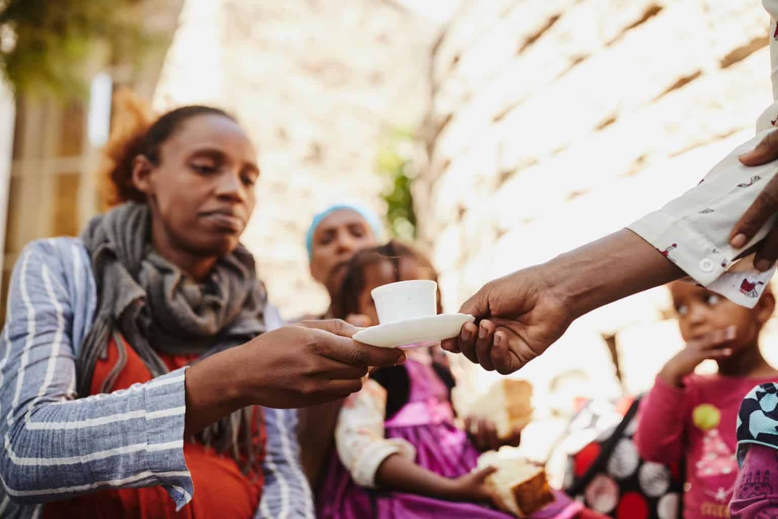 A woman receives a small cup and saucer from another person outdoors; two children and another adult sit nearby, one child wearing a pink dress. The scene suggests a communal or family gathering.