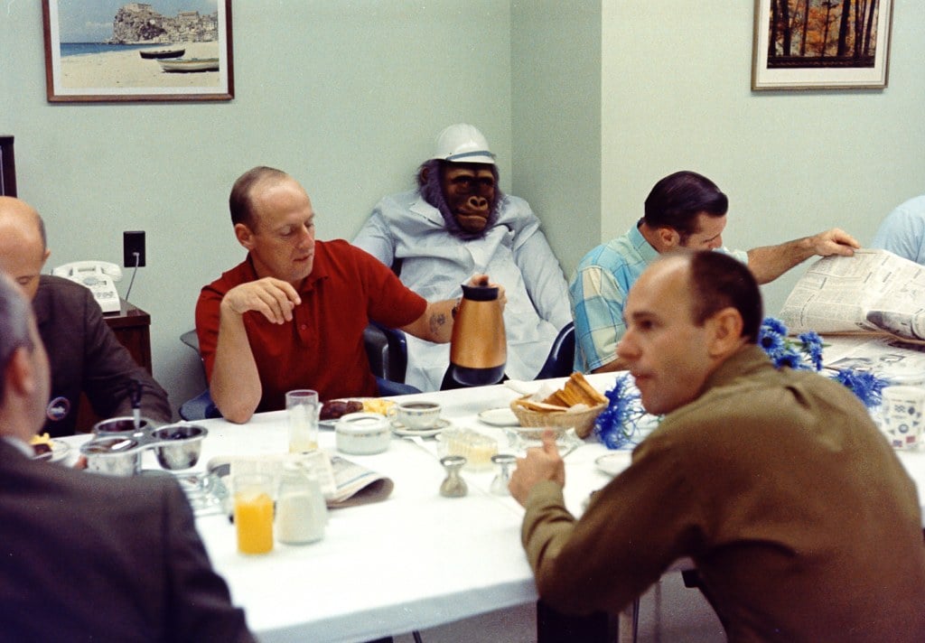 Several men sit around a table having a meal, with one person in the center dressed as a chimpanzee in a white lab coat and cap. The table is set with food, drinks, and newspapers in a casual setting.