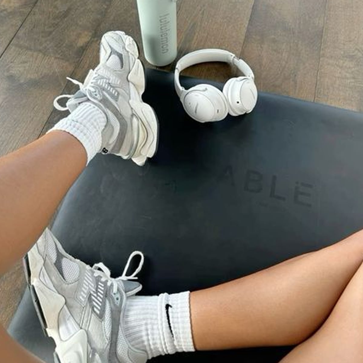 Person wearing white sneakers and Nike socks sits on a black exercise mat, with a pair of white headphones and a light-colored water bottle placed nearby on a wooden floor.