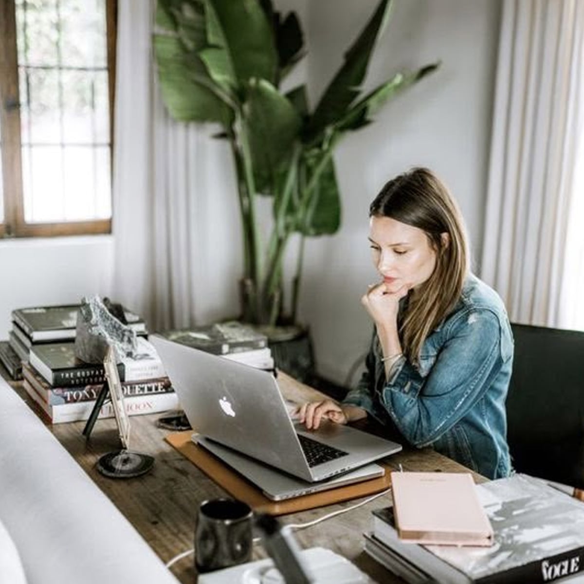 A woman in a denim jacket works at a wooden desk with a laptop, surrounded by books, a notebook, and a mug. Indoor plants and natural light from a nearby window create a cozy workspace atmosphere.