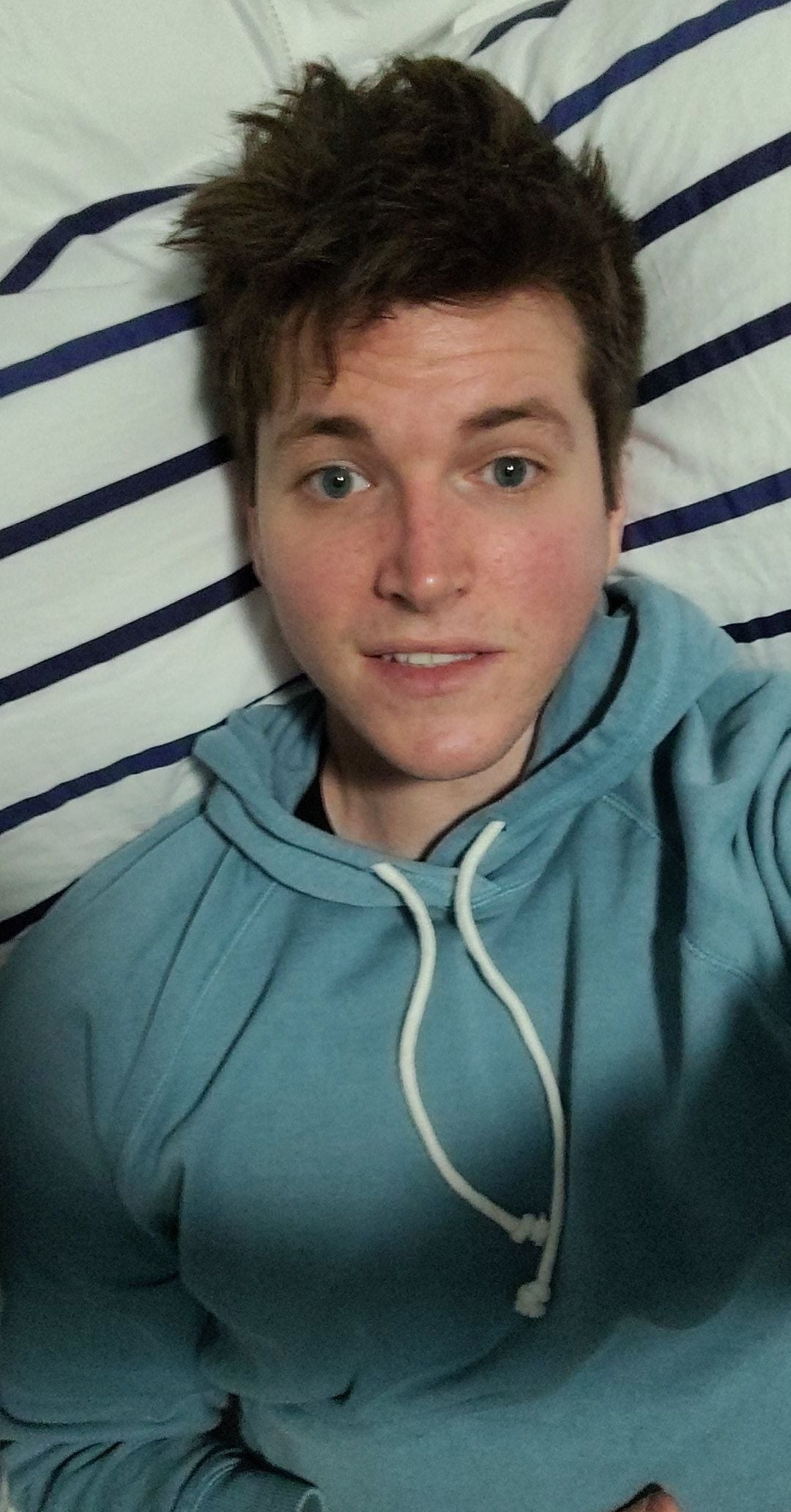 A young person with tousled brown hair and blue eyes is lying on a bed with white and navy striped sheets, wearing a light blue hoodie and looking up at the camera with a slight smile.