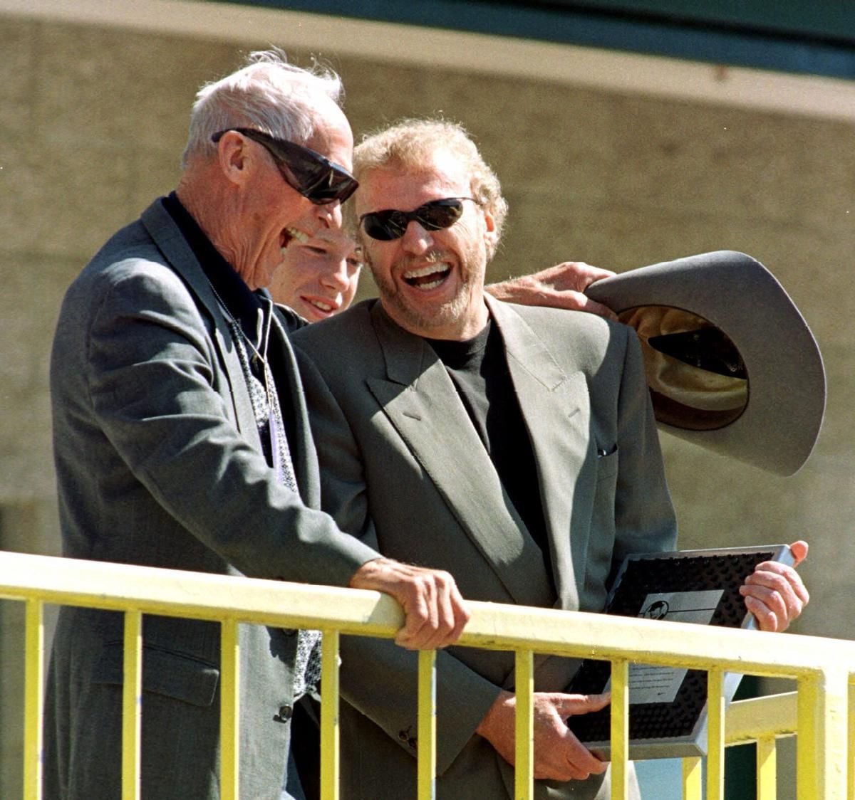 Three men in suits and sunglasses stand on a yellow railing outdoors, smiling and laughing. One man holds a framed plaque while another holds a large hat, partially obscuring his face.