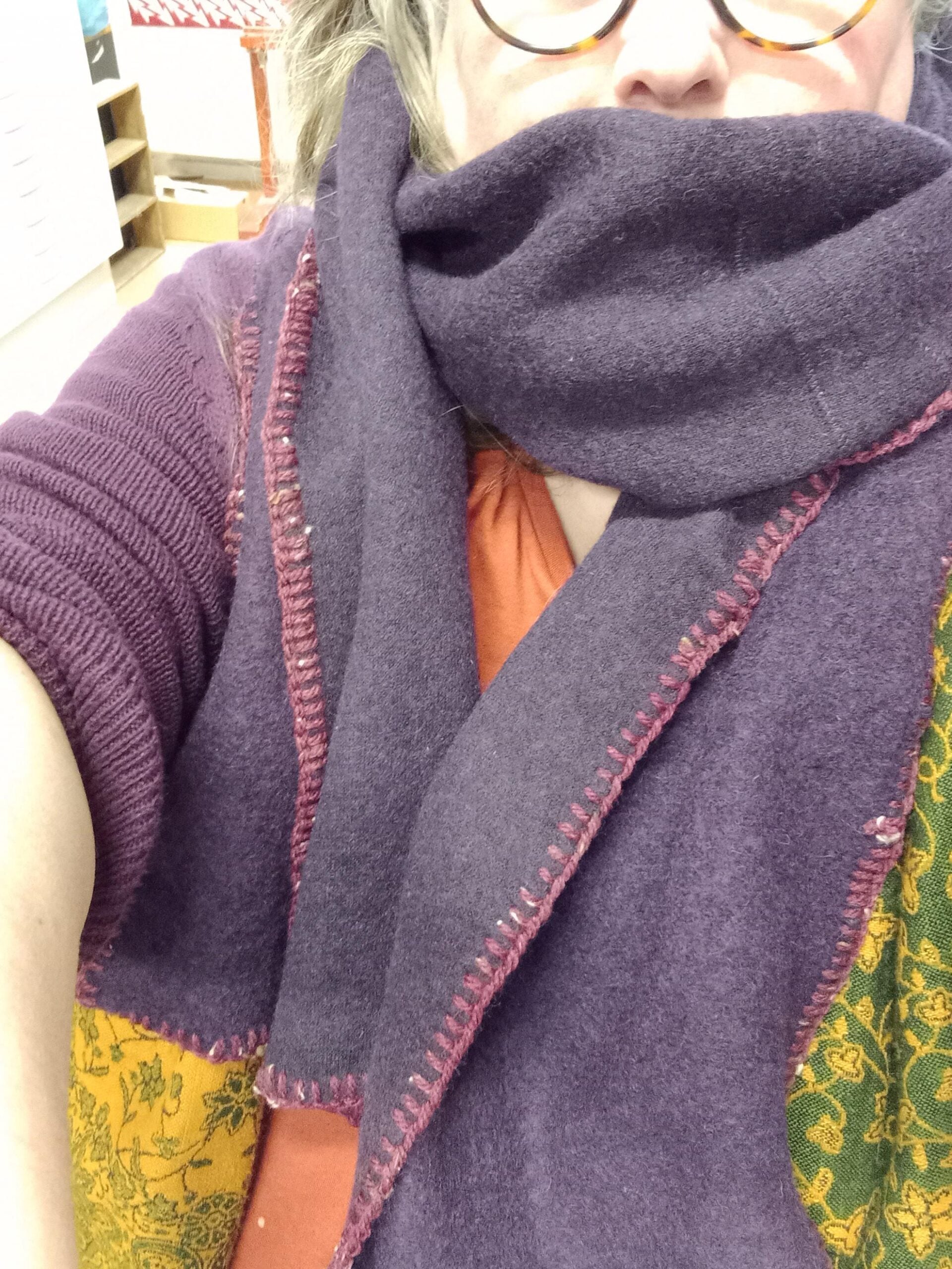 A person wearing glasses, an orange shirt, and a large purple scarf with pink stitching. The scarf covers their mouth and neck, and part of their sweater has green and yellow patterns. The background appears to be indoors.