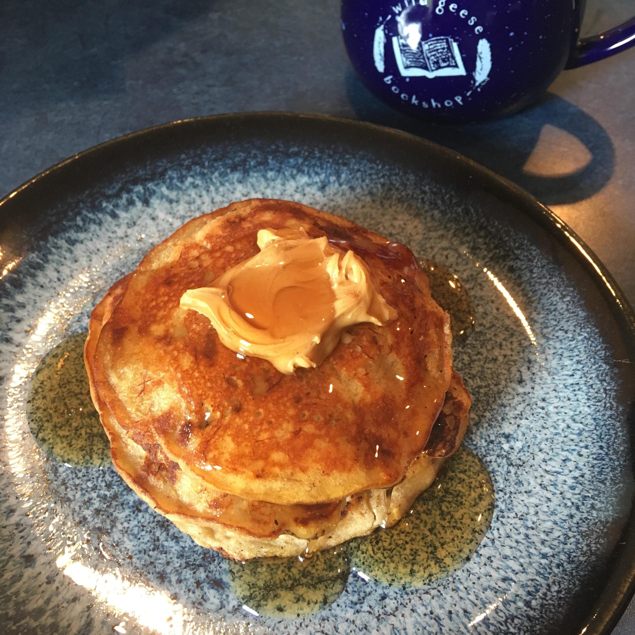 A stack of pancakes topped with a dollop of butter and drizzled with syrup sits on a blue plate; a blue mug with white text and a goose graphic is in the background.