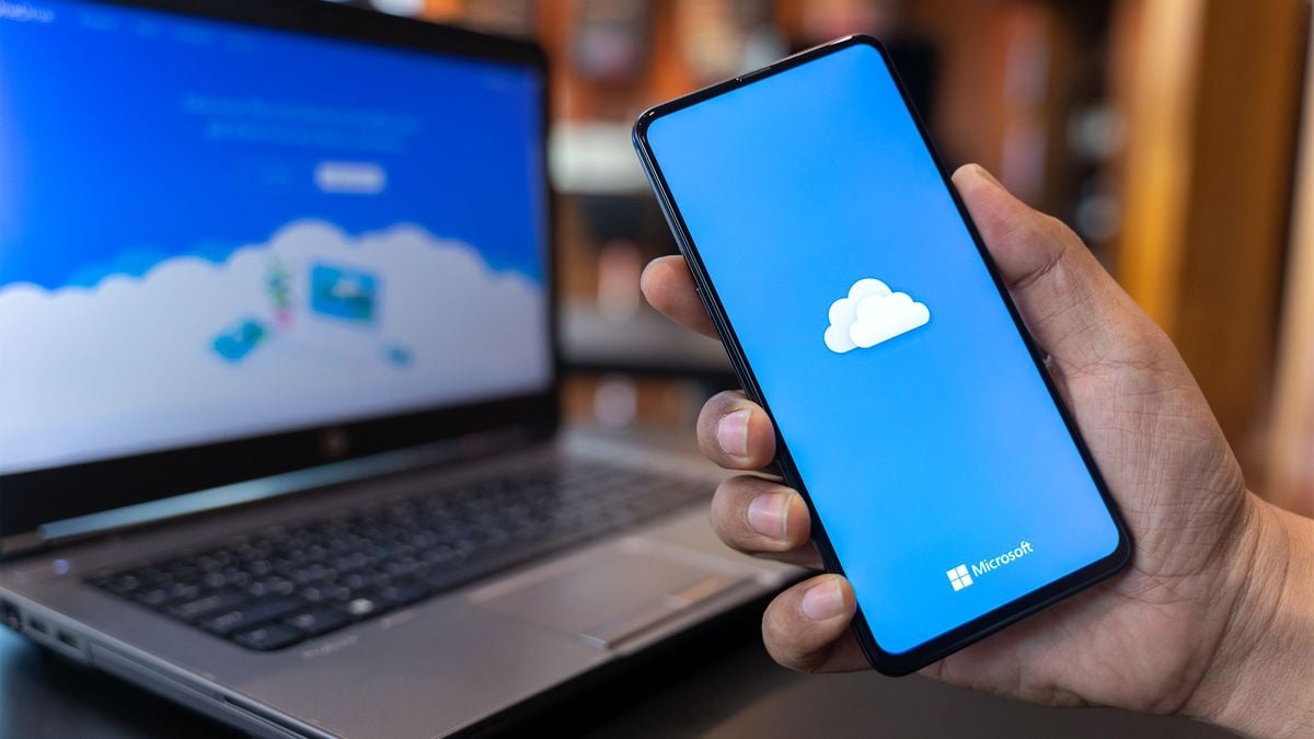 A hand holds a smartphone displaying the Microsoft OneDrive cloud logo, with a laptop showing a blue cloud-themed webpage in the background.