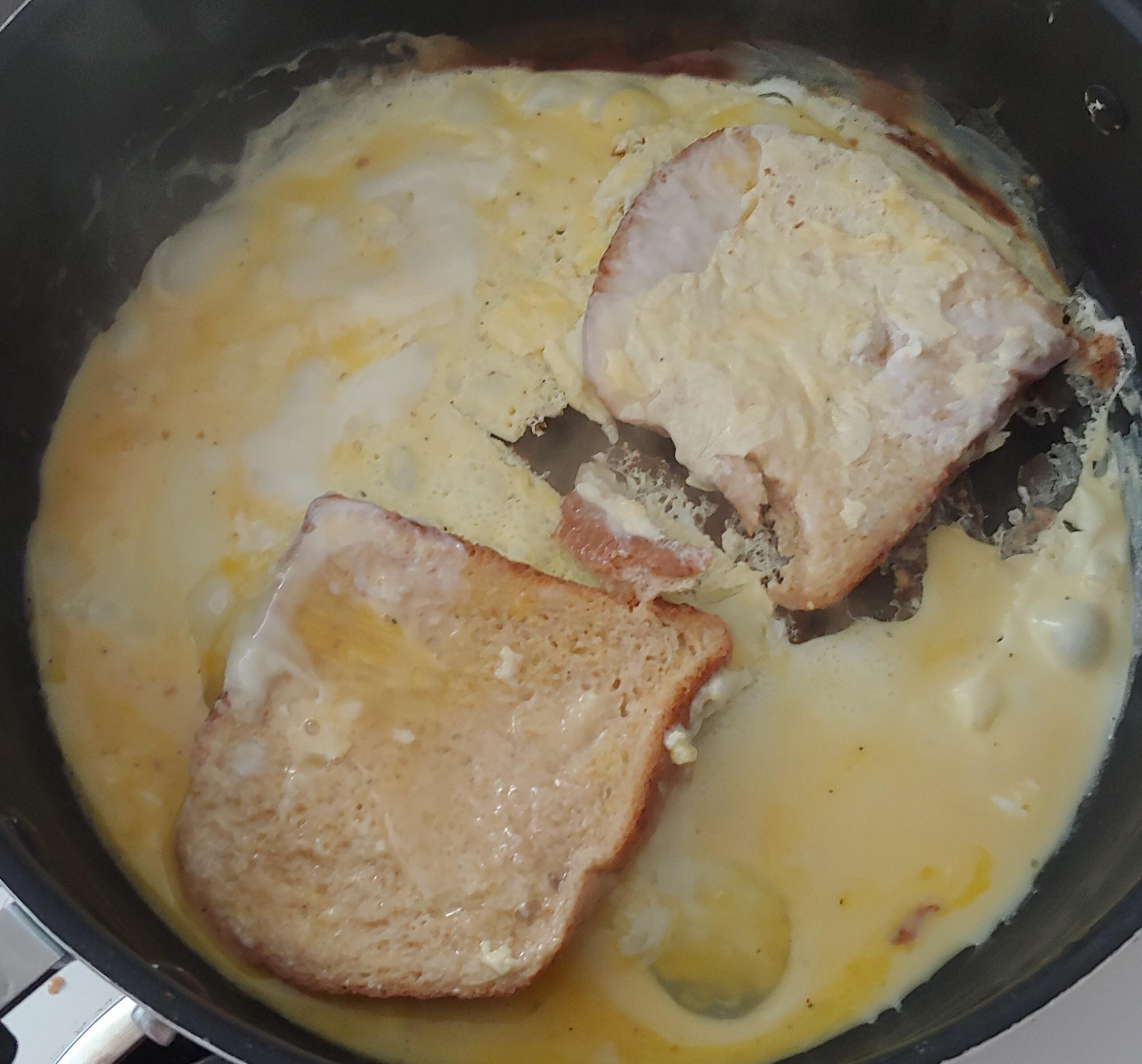 Two slices of bread soaking in a mixture of beaten eggs that are partially cooked in a frying pan, with some egg starting to set around the edges.