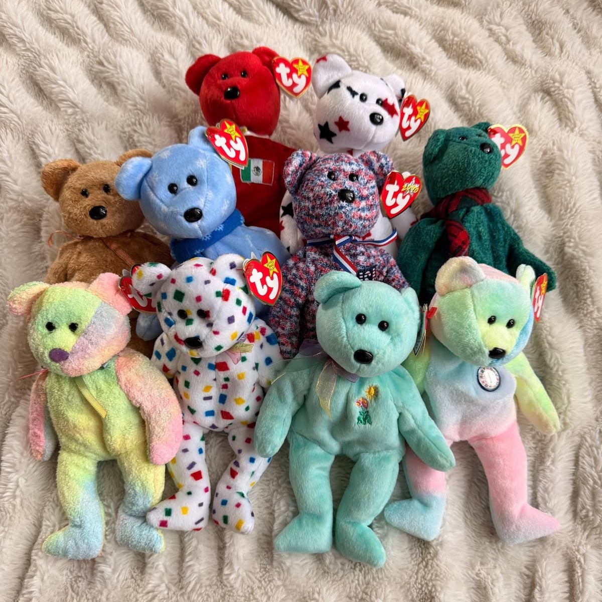 Nine colorful Ty Beanie Baby bears with tags are arranged on a soft, cream-colored textured surface. The bears are in various colors and patterns, including solid, pastel, and multi-colored designs.