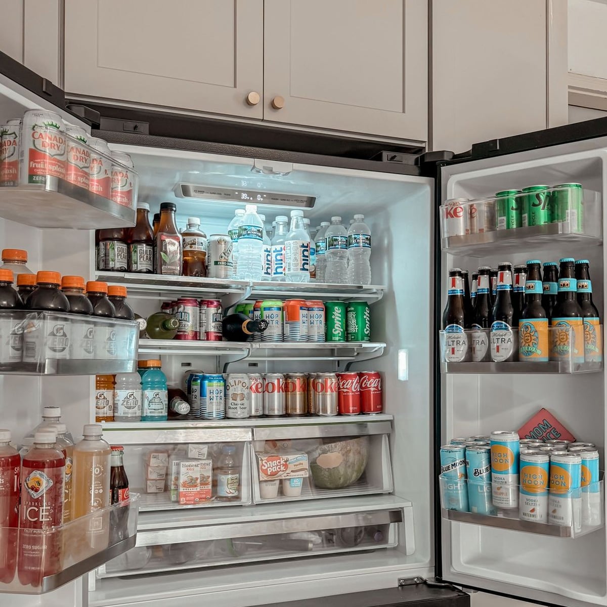 An open refrigerator filled with various beverages, including soda cans, bottled water, sports drinks, iced teas, and beer bottles. The fridge is neatly organized, with drinks arranged on shelves and in the door compartments.