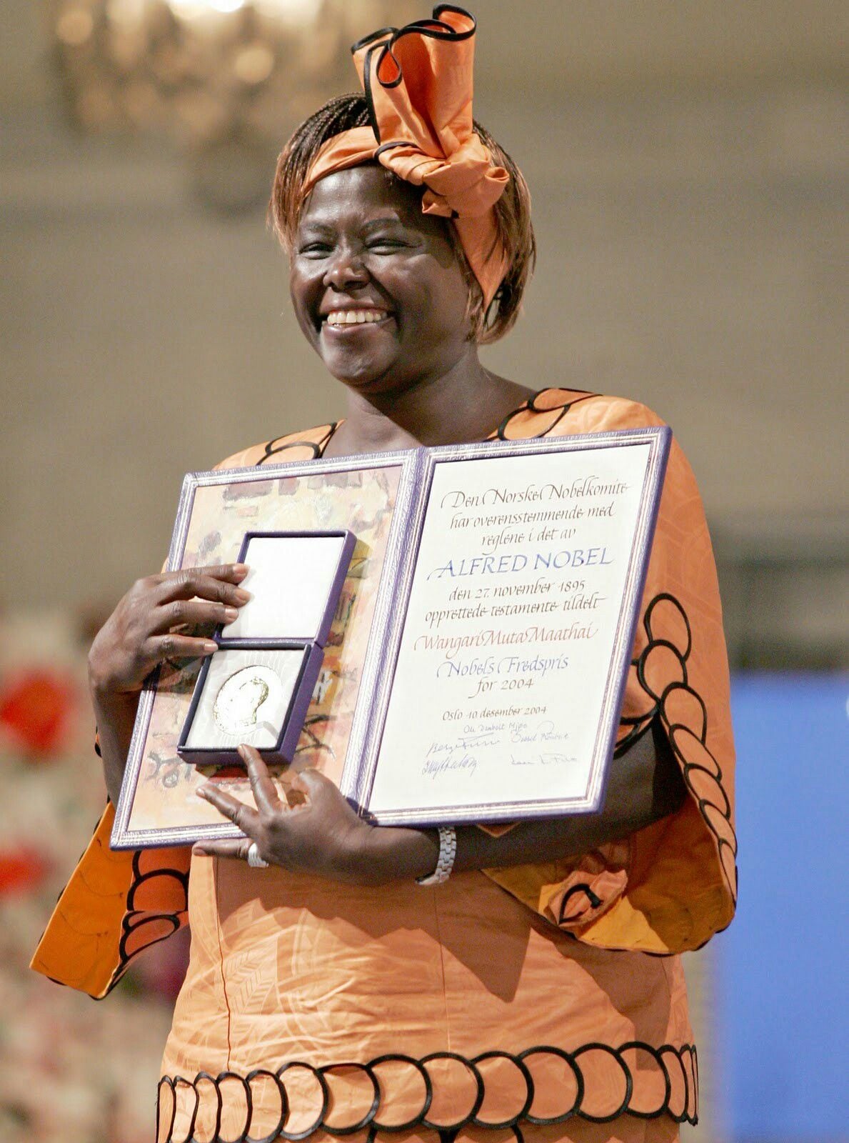 A smiling woman wearing a patterned orange dress and headwrap holds up her Nobel Peace Prize medal and certificate on stage.