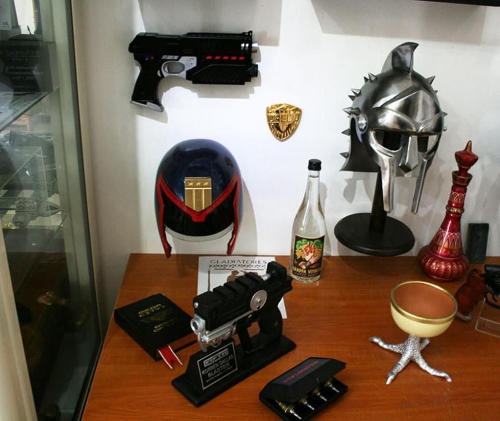 A desk displays sci-fi and fantasy memorabilia, including two futuristic guns, a Judge helmet, a gladiator helmet, a sheriff badge, a goblet on a clawed base, a bottle with a vintage label, and a red decorative piece.
