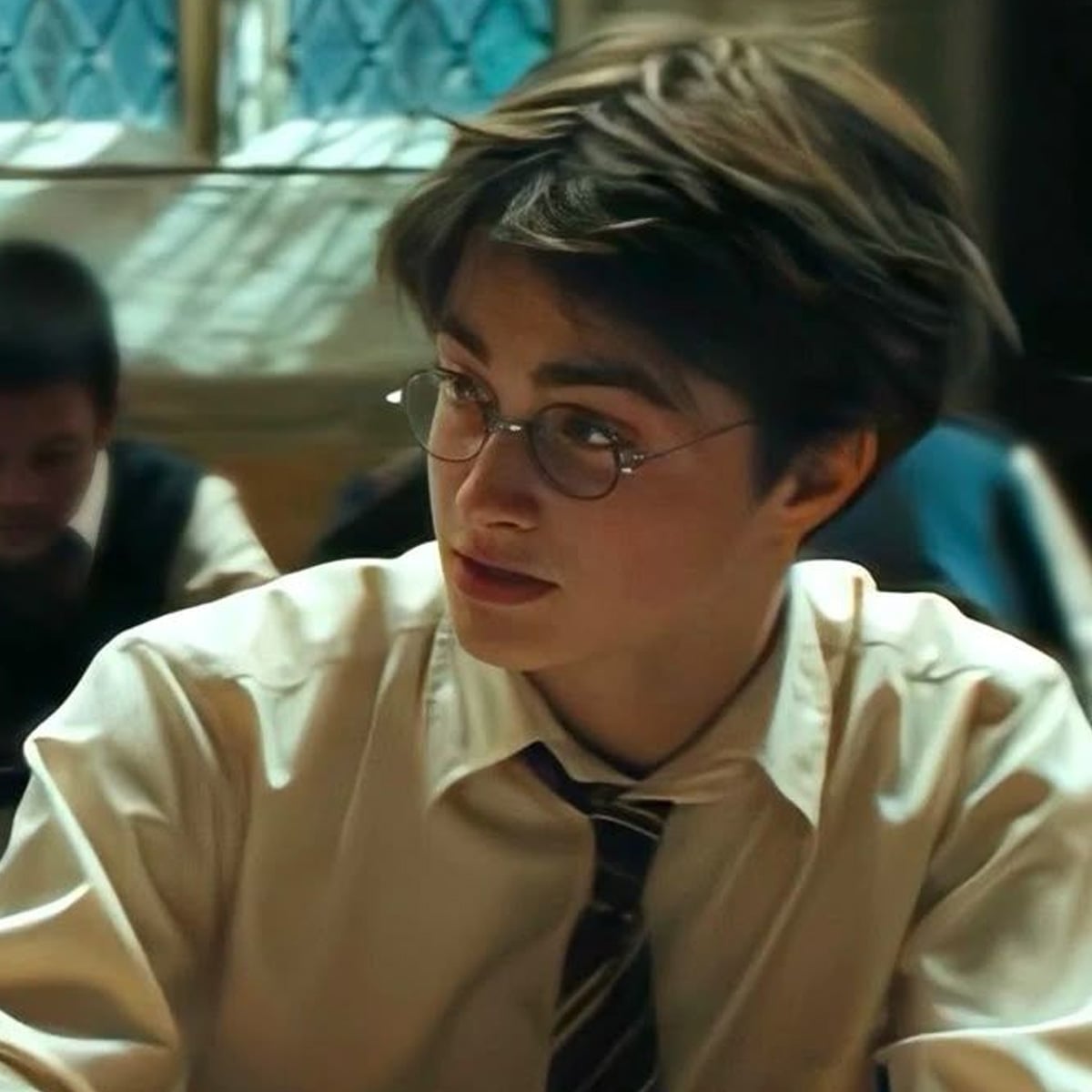 A young person with round glasses, tousled brown hair, and a striped tie sits at a desk in a classroom, looking to the side. The background is blurred, showing another student and a window with diamond-patterned panes.
