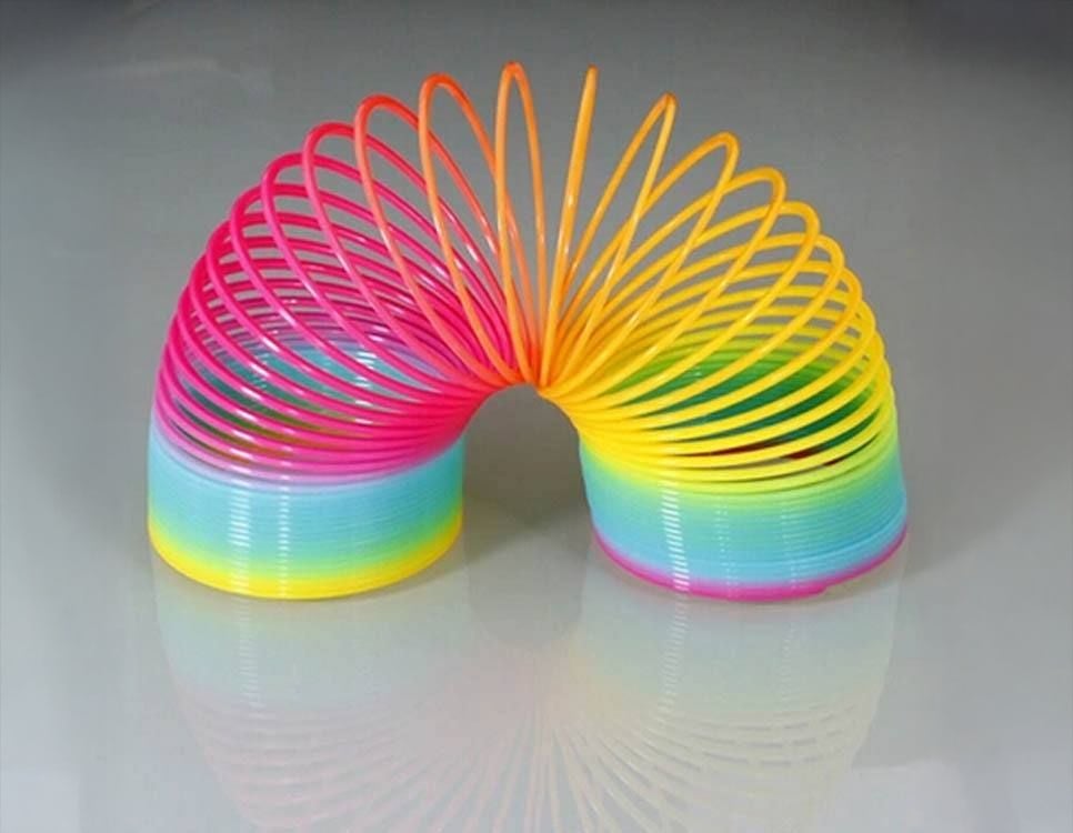 A colorful plastic Slinky toy in a rainbow gradient of pink, yellow, green, and blue, stretched into an arch shape on a reflective surface.